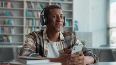 Kulaklık takan Afrikalı Amerikalı adam cep telefonu akıllı telefon öğrenci kütüphanesi, üniversite teknolojisi gülümseyen lise öğrenimi kitapçı etnik kitapçı erkek adam rahatla
