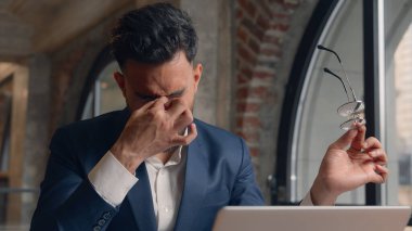 Arap Hintli iş adamı erkek işadamı ofiste dizüstü bilgisayar başında çalışan erkek baş ağrısı çekiyor migren gözlüğü çıkarıyor kaşınan gözler gergin bulanık görme yorgunluğu yorgun aşırı çalışma sağlık sorunu