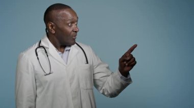 Beyaz önlüklü Afrikalı doktor doktor erkek sağlık çalışanı stüdyo mavi arka planda sağlıkçı müşteri işaret parmağı ile gülümseyerek iyi bir sağlık sigortası seçmeyi seçiyor.