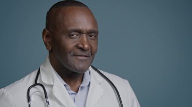 Yakın plan başarılı gülümseyen Afro-Amerikan erkek doktor sağlık çalışanı, portre uzmanı, kardiyolog, pediatrist dişçi, kamera görüntüsü mavi arka plan stüdyosunda.