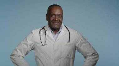 Gülümseyen mutlu Afro-Amerikan erkek profesyonel doktor erkek doktor başarılı etnik çalışan klinik cerrahı stüdyonun mavi arka planında kamera sağlık sigortasına bakıyor.