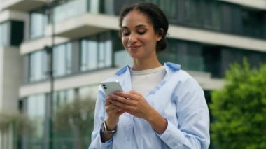 Neşeli, gülümseyen iş kadını Afrikalı Amerikalı iş kadını cep telefonuyla sohbet ediyor internet üzerinden akıllı telefon mesajları alıyor şehir dışında çalışan yönetici kız işveren kameraya gülümsüyor.