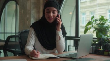 Kadın danışman mutlu gülümseme Arap Müslüman kadın tesettüre tesettürlü iş kadını iş kadını iş kadını cep telefonu konuşması çok görevli yönetici cep telefonu konuşması ofis bilgisayarında dizüstü bilgisayarı çalışma notları yaz