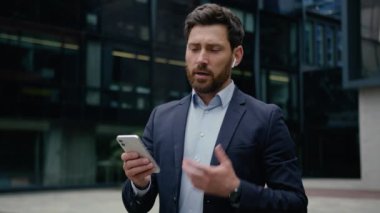 Kafkasyalı iş adamı yetişkin girişimci erkek kablosuz kulaklıklı erkek iş danışmanı işveren cep telefonuna cevap veriyor. Açık havada kullandığımız cihazlarla cep telefonuyla.