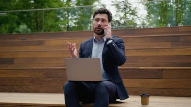 Çok yönlü iş adamı Kafkasyalı iş adamı erkek girişimci şehirli iş adamı şehirde oturup bilgisayarla gülen cep telefonlu akıllı telefon açık havada internet bağlantısı yazan internet bilgisayarı.