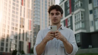 Kafkasyalı adam elinde cep telefonu, akıllı cep telefonu mesajlaşması çevrimiçi sosyal medya sohbetleri, gülümseyen erkek kullanıcı işadamı, kullanıcı iş adamı şehir dışından işçi internet zımbırtısı iş dünyası gökdelenleri.