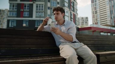 Kafkasyalı bir adam bankta oturuyor, cep telefonu kullanıyor, akıllı telefon kullanıyor, sosyal medya sohbet ediyor, kahve içiyor, gülümseyen şehir işadamı, elinde kupa internet ağı işi tutan işadamı dışarıda.