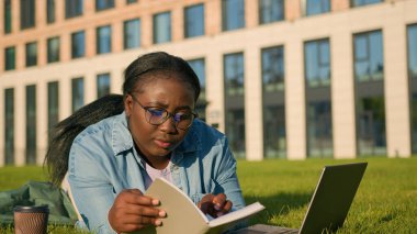 Afro-Amerikan mutlu kadın üniversite akademisi genç kız lisesi parkta yeşil çimlere uzanmış kitap telif defteri bilgisayarlarıyla çalışıyor sınav sınavlarına hazırlanmak için kahve içiyor