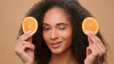 Close up ethnic model holding two slices of orange citrus posing beige background African American girl woman vitamin diet nutrition cosmetology skin care haircare moisturizing oil serum facial cream clipart