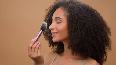 Kapalı portre. Elinde fırça tozu tutan Afrikalı Amerikalı etnik kadın modeli. Bej stüdyo arka planında kameraya gülümseyen cilt tozu sür.