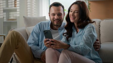 Millennial couple Caucasian man woman married husband wife at home talking using smartphone together happy family discuss offers choosing shopping online with mobile phone boyfriend showing thumb up