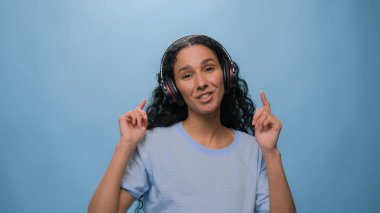 Stüdyo portresi mutlu, gülümseyen Arap kadını söylüyor, Hintli bayan İspanyol, kulaklıkla müzik dinliyor, dans ediyor, radyo dansı yapıyor, mavi arka planda, eğlenceli bir müzik ritmi var.