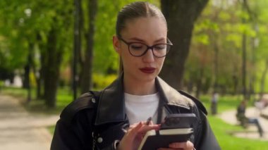 Beyaz modaya uygun, kendine güvenen bir kadın park kullanıcısı cep telefonu akıllı cep telefonu sanal mesajlar mesajlaşması şehir dışında çalışan iş kadını yürüyüş kadın teknolojisi