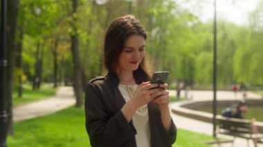 Düşünceli bir kadın parkta cep telefonuyla yürüyor akıllı telefon cihazıyla mesajlaşıyor çevrimiçi sohbet bağlantıları gülümsüyor mutlu mesajlar düşünüyor teknoloji web siteleri sosyal ağları dışarıda gezen bir kız dışında şehri geziyor.