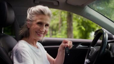 Mutlu gülümseyen yaşlı, olgun, beyaz, kadın, kadın, araba sahibi, kiralama servisi müşterisi. Otomobil anahtarlarını içeriden aldığını gösteren kamera dostu gülümsemeye bakıyor.