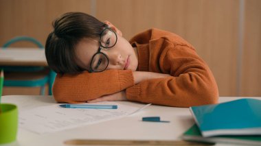Yorgun, bitkin, yorgun, üzgün, üzgün, yorgun, küçük çocuk çocuk, gözlüklü, ilkokul çocuğu okul masasında yatıyor.