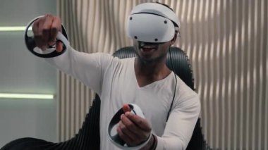 Fütürizm oyun interneti video oyunu sanal gerçekliği VR gözlüklü Afrikalı Amerikalı erkek adam beyaz fütüristik odada 3 boyutlu dünya oynuyor kapalı mekanlarda modern internette eğleniyor metaverse oynamaktan zevk alıyor.