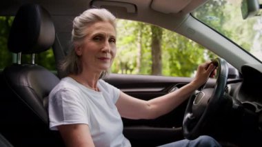 Mutlu gülümseyen Kafkasyalı olgun yaşlı kadın araba sahibi yaşlı kadın büyük anne otomobil şoförü yeni modern elektrikli arabanın içinde oturuyor direksiyon başında kamera görünümlü emeklilik aracı sürüyor.