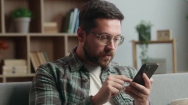 Evde şaşkın şaşkın şaşkın şaşırmış cihaz bağımlısı erkek cep telefonu uygulaması, akıllı telefon sosyal medya, kötü haber, internet sitesi, kanepe üzerinde rahatla.