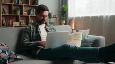 Mutlu, gülümseyen, beyaz, erkek, kanepesinde dizüstü bilgisayarı olan, internet üzerinde çalışan, alışveriş yapan, işini yapan, rahatlayan, ellerini başının arkasına koy.