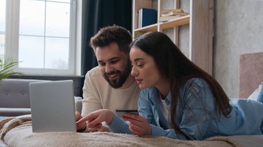 Kafkasyalı çift yatakta yan yana yatıyorlar kredi kartı kullanıyorlar dizüstü bilgisayar bankacılığı uygulamasına şifreyi giriyorlar gülümseyen kadın ve erkek alışveriş siparişleri ev yatak odasında ödeniyor.