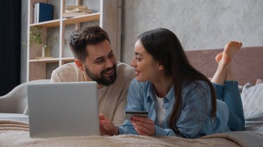 Kafkasyalı çift yatakta yan yana yatıyorlar kredi kartı kullanıyorlar dizüstü bilgisayar bankacılığı uygulamasına şifreyi giriyorlar gülümseyen kadın ve erkek alışveriş siparişleri ev yatak odasında ödeniyor.
