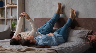 Kafkasyalı mutlu çift, rahat bir yatakta baş aşağı uzanıyorlar. Cep telefonlarıyla internet üzerinden alışveriş yapıyorlar. Kadın ve erkek kocaları yatak odasında dinleniyorlar.