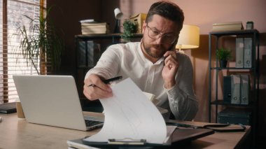 Kafkasyalı orta yaşlı bir adam cep telefonuyla konuşuyor ofis işlerinde çoklu iş görüşmesi erkek şirket patronu iş adamı tüccarlarına bilgisayar kağıtlarıyla yürütme planları yapıyor.