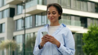 Tatmin olmuş Afro-Amerikan kadın kadın kadın iş kadını kadın kullanıcı cep telefonu kullanıcısı elinde taksi siparişi internet uygulaması akıllı telefonu açık havada kameraya bakıp gülümsüyor.