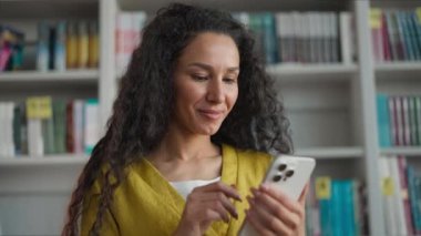 Heyecanlı Latin Kafkasyalı kadın elinde cep telefonu, akıllı telefon, gülücük, duygusal sınav sonuçları lise kütüphanesindeki kız öğrenci, sosyal medya çalışmalarından keyif alıyor.