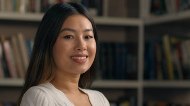 Yakın plan gülümseyen Asyalı kadın kapalı mekanda kameraya dönüyor mutlu Çinli etnik kız öğrenci iş kadını ofis çalışanı kitaplığın yanında kadın portresinin yanında poz veriyor.
