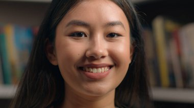 Yakın plan gülümseyen Asyalı kadın kapalı mekanda kameraya dönüyor mutlu Çinli etnik kız öğrenci iş kadını ofis çalışanı kitaplığın yanında kadın portresinin yanında poz veriyor.