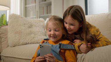 İki neşeli Kafkas çocuk kız arkadaş çocuklar oturma odasındaki kanepede oyun oynayarak eğleniyorlar internetteki akıllı telefon sosyal ağını kullanarak.