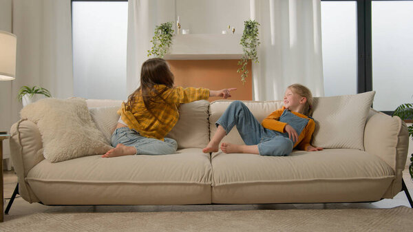 Two laughing little Caucasian children friends kids girls playing couch pointing finger gesture you sisters relationship daughters family fun talking contend debate sharing sofa together living room