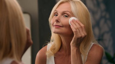 50 'li yaşlarda, güzel, olgun bir kadın yüzüne pamuk yastıkla toz ve lüks kozmetik uyguluyor. Kusursuz pürüzsüz cilt makyajı yapıyor. Aynalı ev banyosunda, yüz kırışıklıkları, yaşlanmayı önleyen güzellikler.