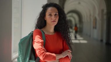 Kafkasyalı, ciddi, barışçıl, Latin kökenli, Latin kökenli, bayan öğrenci üniversite akademisi koridorunda eğitim öğrenim görüyor ve kamera fotoğraflarına bakıyor.