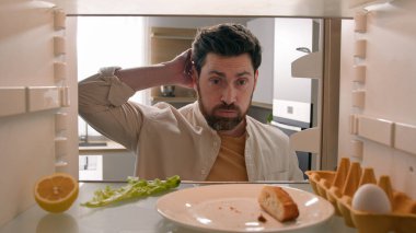 Hungry man guy open empty refrigerator with half lemon lettuce leaf one egg slice of bread leftover orange juice in bottle confused puzzled sad need food delivery point of view POV from inside fridge