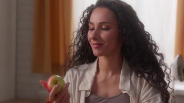Mutlu İspanyol bayan bayan bayan bayan sağlıklı beslenme yiyeceği yiyor havaya elma fırlatıyor ev mutfağında el ısırığı meyveleri yiyor kameraya gülümsüyor evde sağlık hizmeti ağırlığı kontrolü yiyor