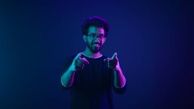 Portrait in neon ultraviolet happy Indian guy Arabian man worker coder hacker computer engineer make gesture hey you showing index finger front direction at camera thumbs up agree support recommend clipart