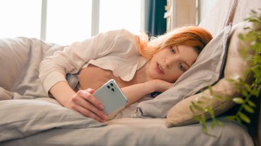 Akıllı telefonlu Kafkasyalı kadını kapat. Sabah yatak odasında uzan, cep telefonlu kadın blogcu kızla sohbet et. Uyandıktan sonra sosyal medyada gülümse.