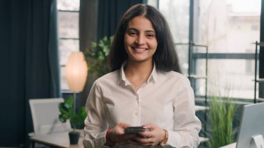 Ofiste cep telefonu kullanan mutlu çok ırklı kadın kameraya gülümseyen Hintli iş kadını Arap kadın yönetici kadın internet sörfçüsü internette geziniyor haber tebessümlerini kontrol ediyor.