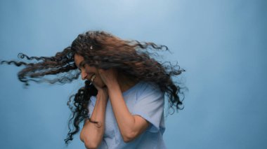 Stüdyo portresi mutlu, gülümseyen Arap kadını söylüyor, Hintli bayan İspanyol, kulaklıkla müzik dinliyor, dans ediyor, radyo dansı yapıyor, mavi arka planda, eğlenceli bir müzik ritmi var.