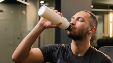 Amerikan çoklu etnik erkek sporcu sporcu sporcu sporcu sporcu sporcu adam spor hocası spor şişesi ile su içen spor salonundaki aktif egzersiz kameraya gülümseyen sağlıklı yaşam tarzına uyum sağlıyor.