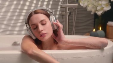 Sakin, sakin, sakin, gözlerini kapatmış, kablosuz kulaklıkla müzik dinleyen, podcast sesi duyan, beyaz, kadın, küvet kaplıcasında dinlenen, sıcak, sıcak su hijyeninden zevk alan bir kadın.