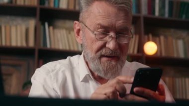Kafkasyalı yaşlı bir adam cep telefonu akıllı telefon zumlayan resim erkek ekran parmakları yaşlı emeklilik işadamı teknolojisi kıdemli internet gezintisi ev içi iş dünyasında olgun görünüm sergiliyor