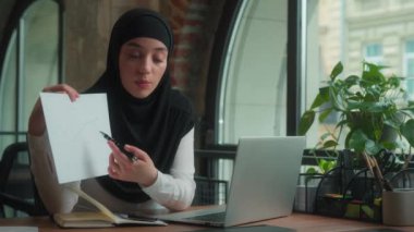Müslüman Arap İslami kız tesettürlü gülümseyen kadın iş kadını kadın dizüstü bilgisayar video görüşmeleri kağıt dökümanına işaret ederek online çatışma rapor satış grafiği iş sunumunu tartıştı