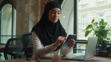 Müslüman kadın tesettüre girmiş Arap iş kadını. Masa başında çalışan gülümseyen iş kadını. Cep telefonu bağımlısı.