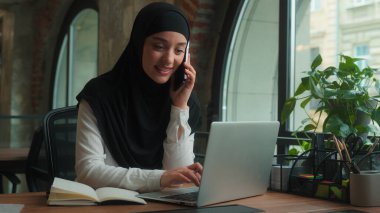 Mutlu Müslüman İslamcı kadın tesettürlü kadın Arap kadın çoklu görev sahibi iş kadını yöneticisi konuşuyor cep telefonu cevapları akıllı telefon görüşmeleri danışma istemcisi ofisteki bilgisayarda yazı yazan notlar yazıyor.