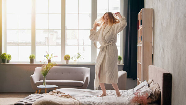 Домашняя утренняя вечеринка в спальне веселая женщина веселится карусель Кавказ счастливая активная девушка в халате танцует прыгая на кровати, двигаясь слушая музыку танцевальный прыжок радостный праздник в роскошном гостиничном номере