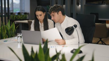 Beyaz kadın ve erkek birlikte çalışıyorlar. Belgeler ve dizüstü bilgisayarla çalışıyorlar. Ofis işleri ile ilgili evrak işleriyle uğraşıyorlar.